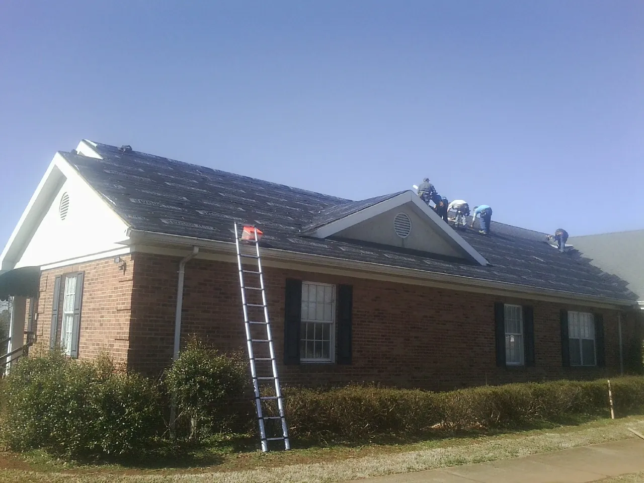 Crew tearing shingles off a brick ranch roof with a ladder set — Metal Roof Repair work by our crew in Kettle Falls