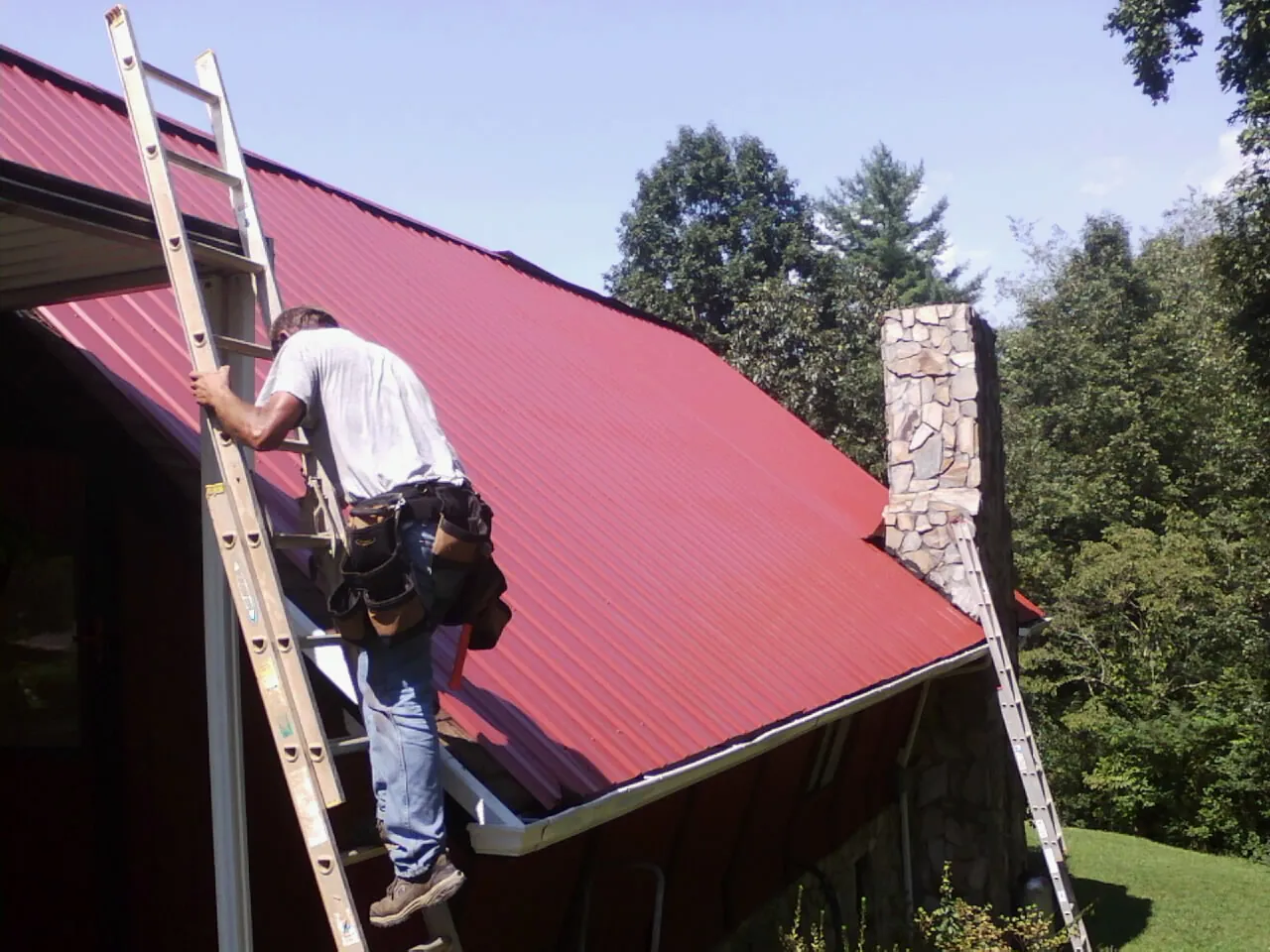 Roofer on a ladder beside a red metal roof and stone chimney — Soffit & Fascia Repair by Roofing Brothers in Kettle Falls